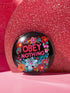 A black round pinback button featuring floral embroidery-style designs and bold red text that reads "Obey Nothing."